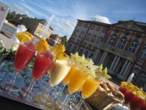Bunte Fruchtcocktails in Gläsern auf einem Tablett vor einem historischen Gebäude mit Säulen und einem Springbrunnen bei sonnigem Himmel