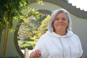 Portrait einer älteren Frau mit kurzen grauen Haaren in weißem Kapuzenpullover vor einem kreisförmigen Torbogen in einem Garten mit Bambuspflanzen und einem weißen Pagoden-Skulptur im Hintergrund
