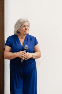 Ältere Frau mit kurzen grauen Haaren in einem blauen Kleid lehnt lächelnd an einer Holzwand und hält ein Mikrofon in beiden Händen, vor hellem Hintergrund.