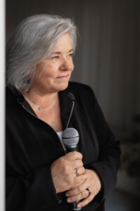 Ältere Frau mit silbernem Haar, die ein Mikrofon hält und nach rechts schaut, trägt schwarzen Reißverschluss-Pullover und Schmuck.