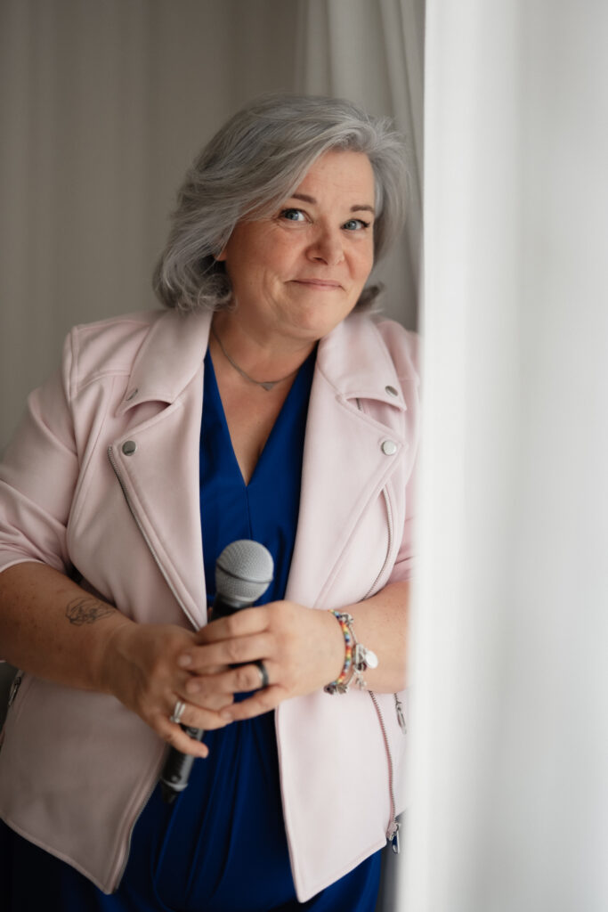 Lächelnde Frau mittleren Alters mit grauen Haaren, die ein blaues Oberteil und eine rosa Jacke trägt, hält ein Mikrofon in beiden Händen und steht neben einem weißen Vorhang in einem hellen Raum.