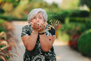 Frau mit weißen Haaren und schwarz-weiß gemustertem Shirt, die bunte Blütenblätter oder Blütenstaub in die Luft vor einem grünen Gartenweg bläst