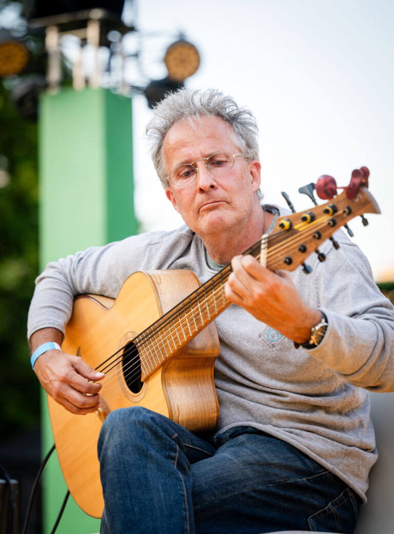 Älterer Mann mit grauem Haar und Brille, der konzentriert eine sechssaitige Akustikgitarre spielt, trägt grauen Pullover und Jeans, Sitz im Freien mit unscharfem Hintergrund.