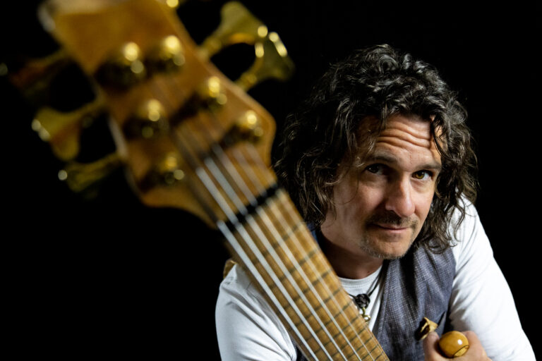 Mann mit lockigem Haar und Schnurrbart, der eine Gitarre hält, vor schwarzem Hintergrund