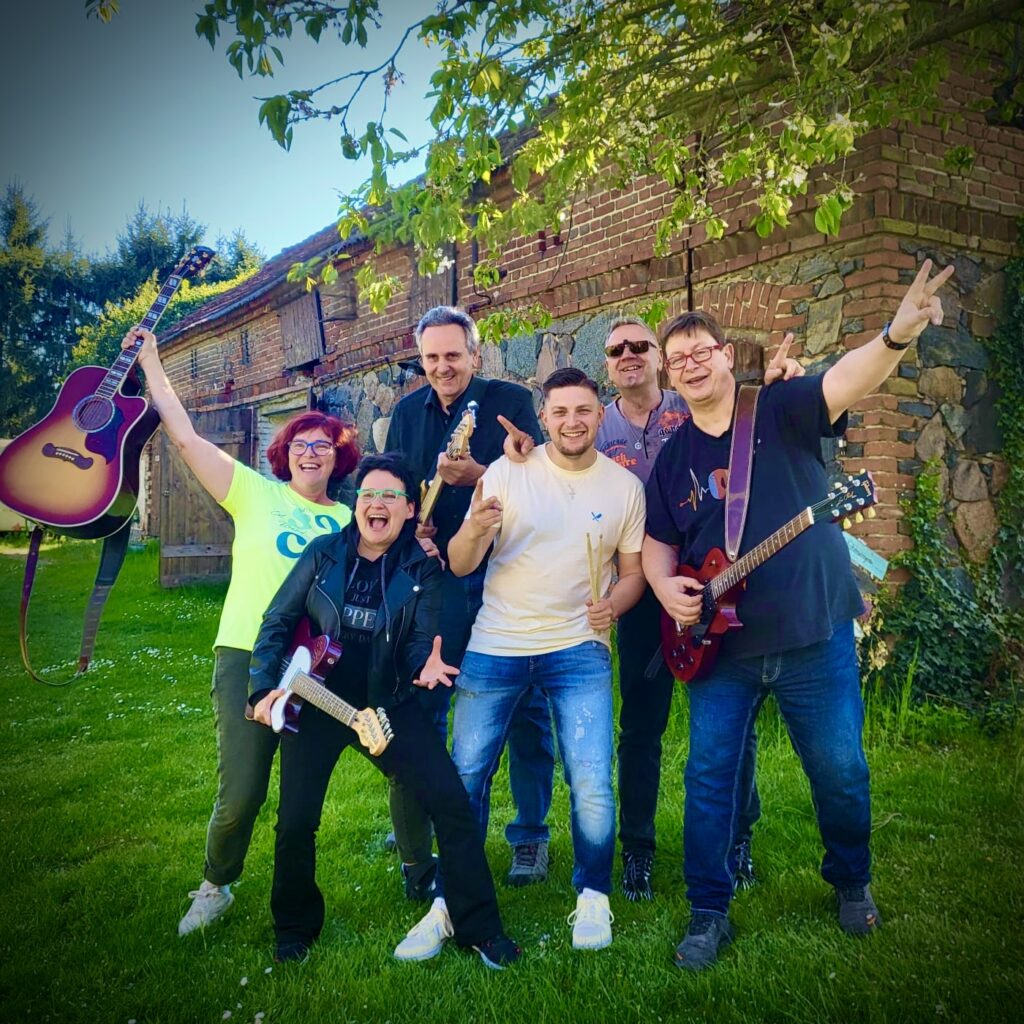 Sechs fröhliche Personen vor einem alten Backsteingebäude mit gründem Rasen, zwei spielen E-Gitarren, eine hält eine Akustikgitarre hoch, eine weitere Person trägt Schlagzeugstöcke, alle posieren ausgelassen und lachen unter einem Baum mit grünen Blättern.