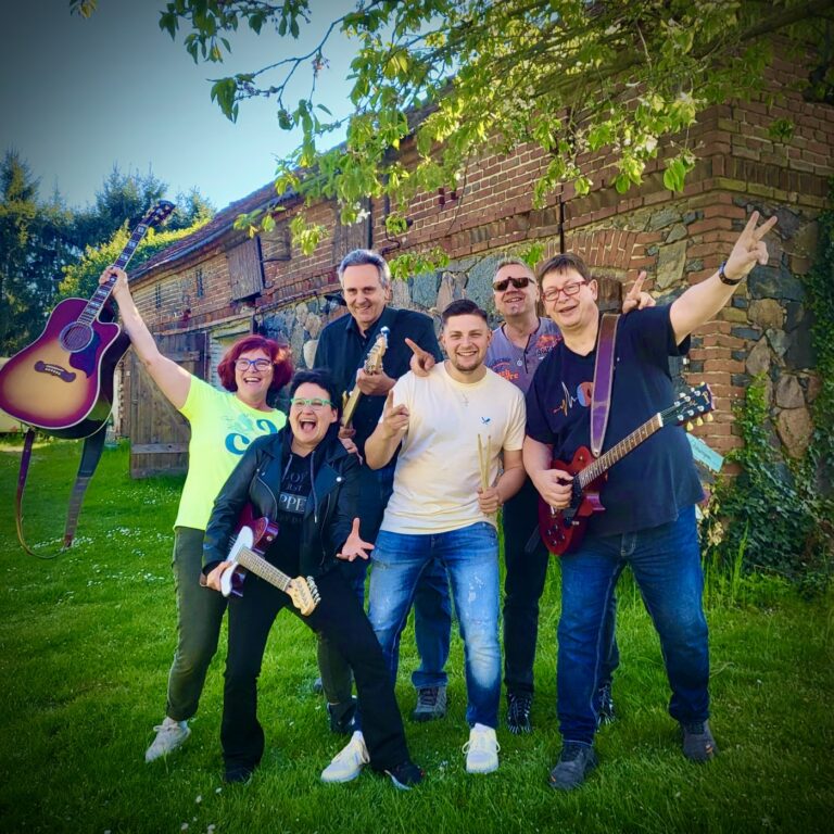 Sechs fröhliche Personen vor einem alten Backsteingebäude mit gründem Rasen, zwei spielen E-Gitarren, eine hält eine Akustikgitarre hoch, eine weitere Person trägt Schlagzeugstöcke, alle posieren ausgelassen und lachen unter einem Baum mit grünen Blättern.