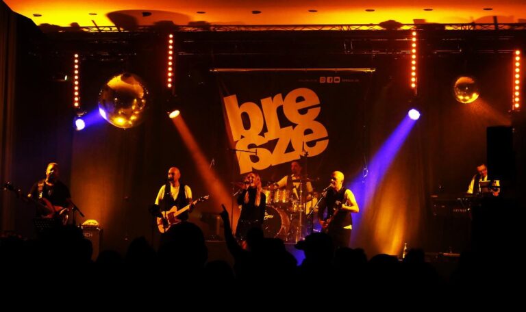 Band »Breshape« spielt live auf einer Bühne mit orangeblauer Beleuchtung, fünf Musiker mit Instrumenten und Sängerin, Publikum als dunkle Silhouetten im Vordergrund