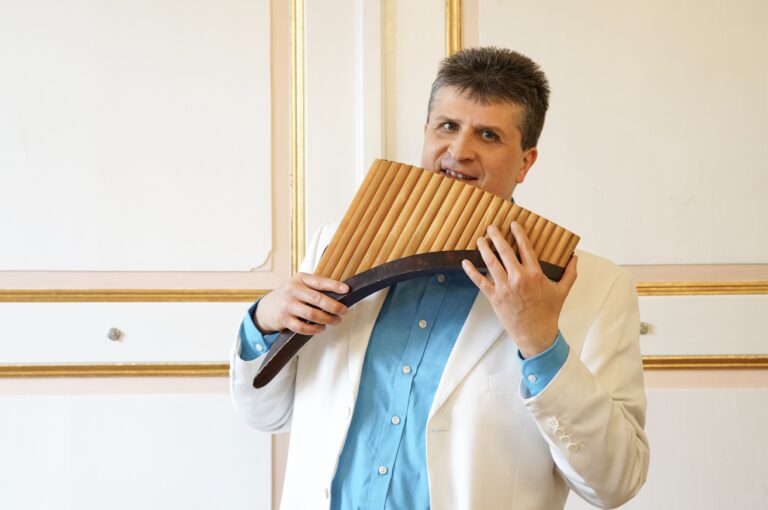 Mann in weißem Sakko und blauem Hemd spielt Panflöte vor einer hellen Wand mit goldgerahmten Paneelen.