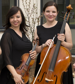 Zwei Frauen in schwarzer Kleidung halten ein Streichinstrument, die linke eine Geige, die rechte ein Cello, im Hintergrund ein Fenster und ein kahler Pflanzenzweig.