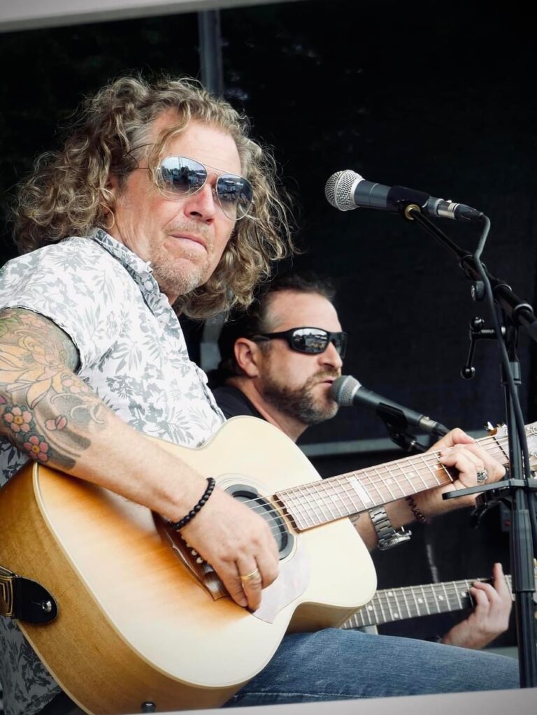 Zwei Männer mit Sonnenbrillen spielen Akustikgitarren und singen in Mikrofone bei einem Auftritt auf einer Bühne.