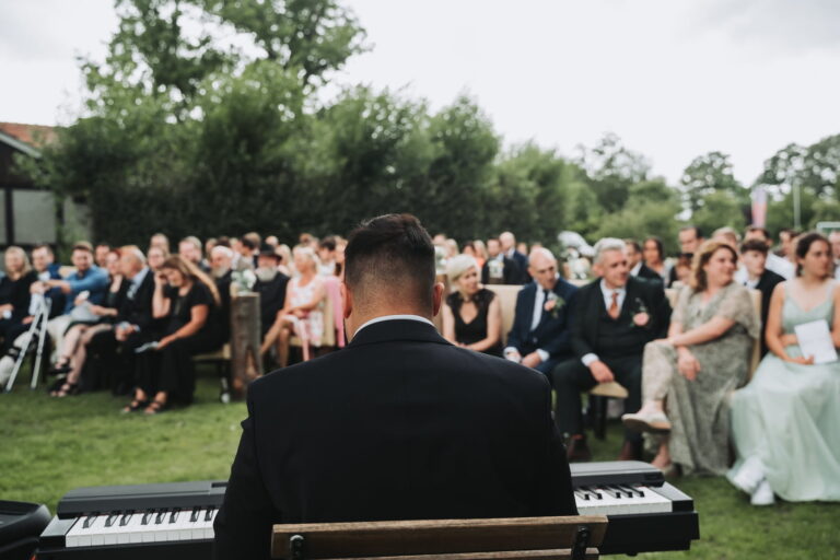 Rückansicht eines Mannes im Anzug, der an einem Keyboard sitzt, umgeben von einer großen Gruppe elegant gekleideter Menschen, die auf einer Wiese mit Bäumen im Hintergrund sitzen und einem Open-Air-Event lauschen.