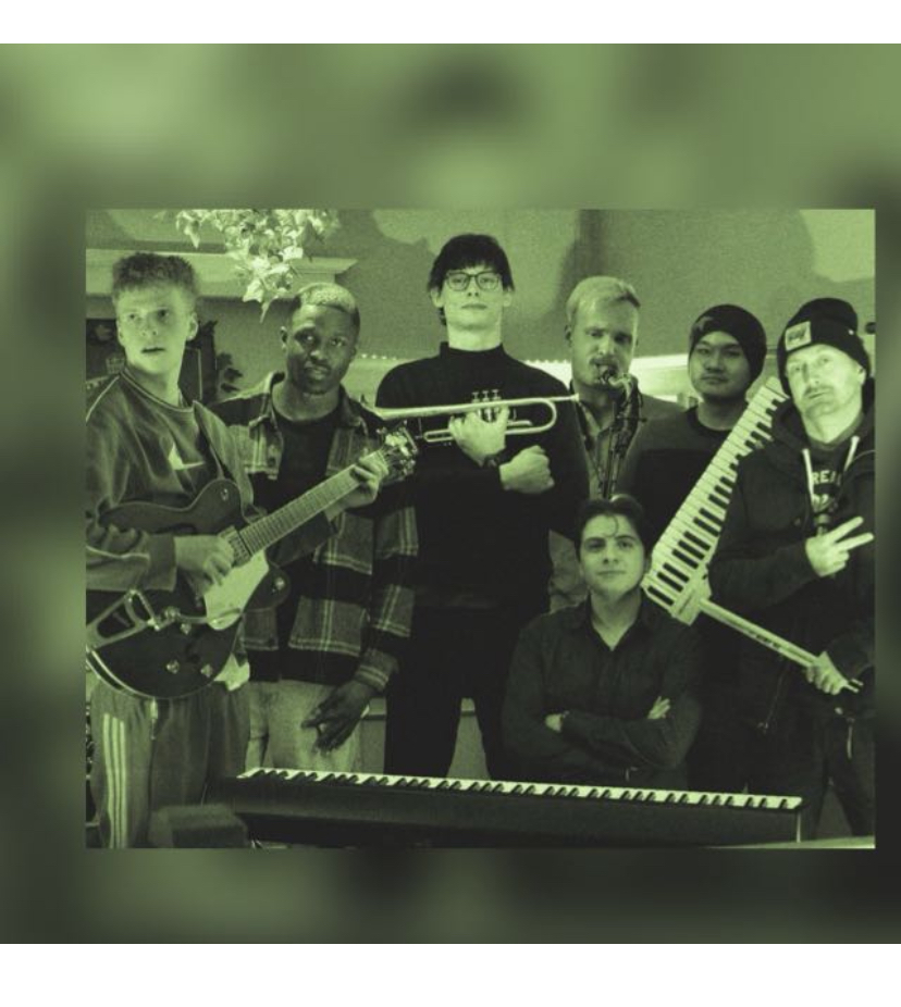 Sieben Männer posieren mit Musikinstrumenten, darunter Gitarre, Trompete, Saxophon, Keytar und Keyboard, vor einem grünen Hintergrund in Innenraum.