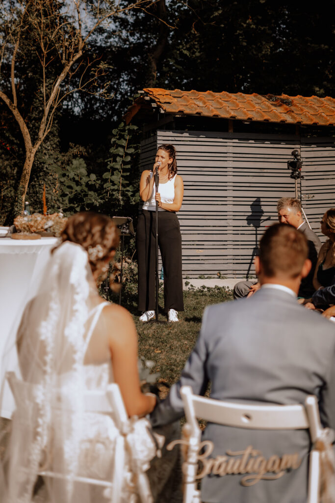 Braut und Bräutigam halten Händchen in weißen Stühlen sitzend mit Blick auf eine Frau, die vor einem Mikrofon bei einer Hochzeit im Freien spricht, mit grünem Hintergrund und Holzhütte mit rotem Ziegeldach.