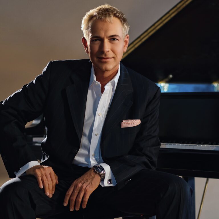 Eleganter Mann mit blondem Haar in dunklem Nadelstreifanzug und weißem Hemd sitzt vor einem schwarzen Klavier und blickt freundlich in die Kamera.