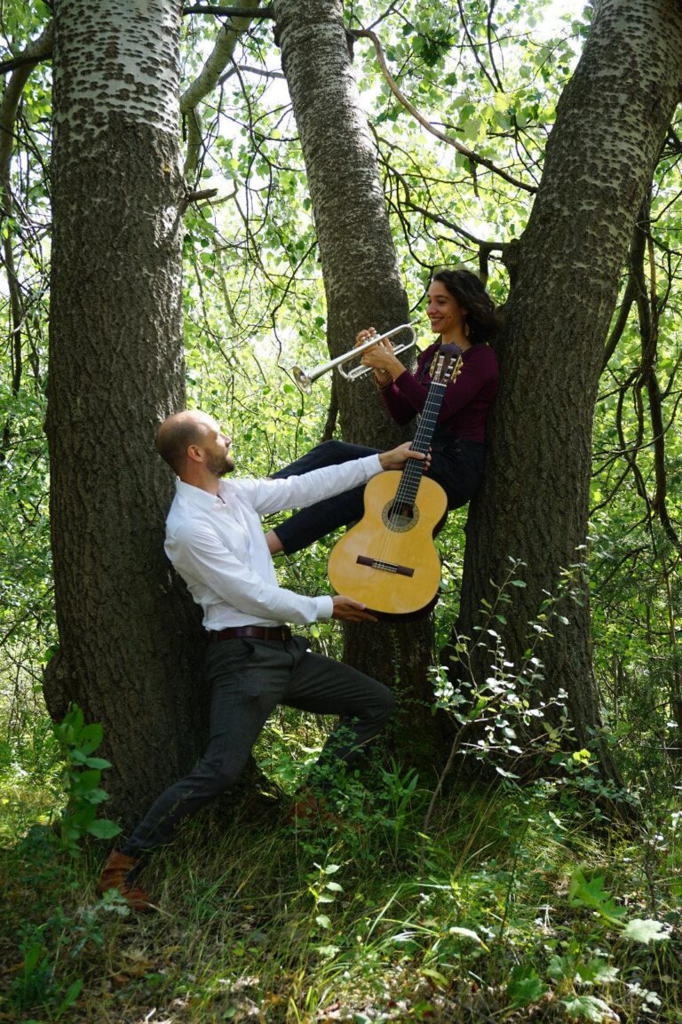 Ein Mann in weißem Hemd und dunkler Hose hält eine Gitarre für eine lächelnde Frau, die in einem Baumsplit sitzt und eine Trompete spielt, umgeben von grünem Laub und Baumstämmen im Wald.