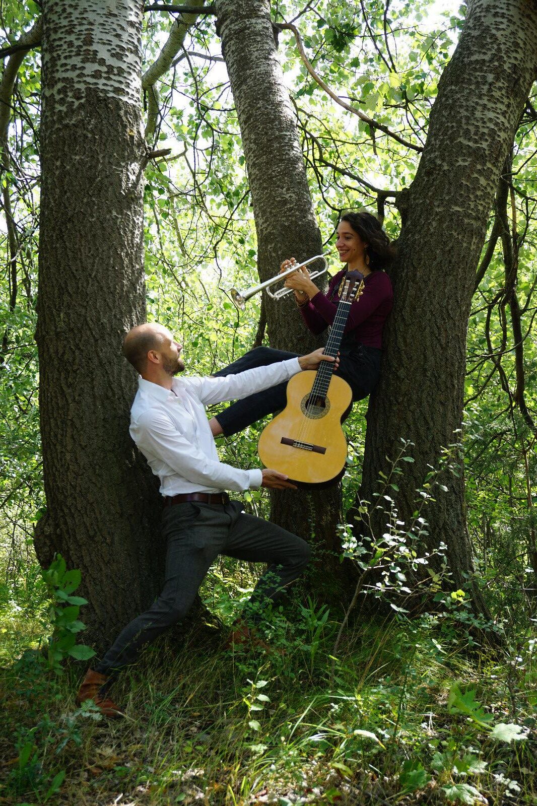 Ein Mann in weißem Hemd und dunkler Hose hält eine Gitarre für eine lächelnde Frau, die in einem Baumsplit sitzt und eine Trompete spielt, umgeben von grünem Laub und Baumstämmen im Wald.