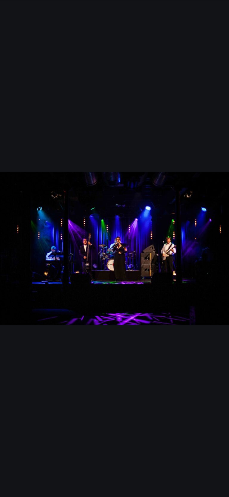 Band auf einer Bühne mit bunter Bühnenbeleuchtung, bestehend aus einem Keyboarder, einem Kontrabass-Spieler, einer Sängerin in schwarzem Kleid und einem Gitarristen, vor einem dunklen Hintergrund mit lila, grünen und blauen Lichtern.