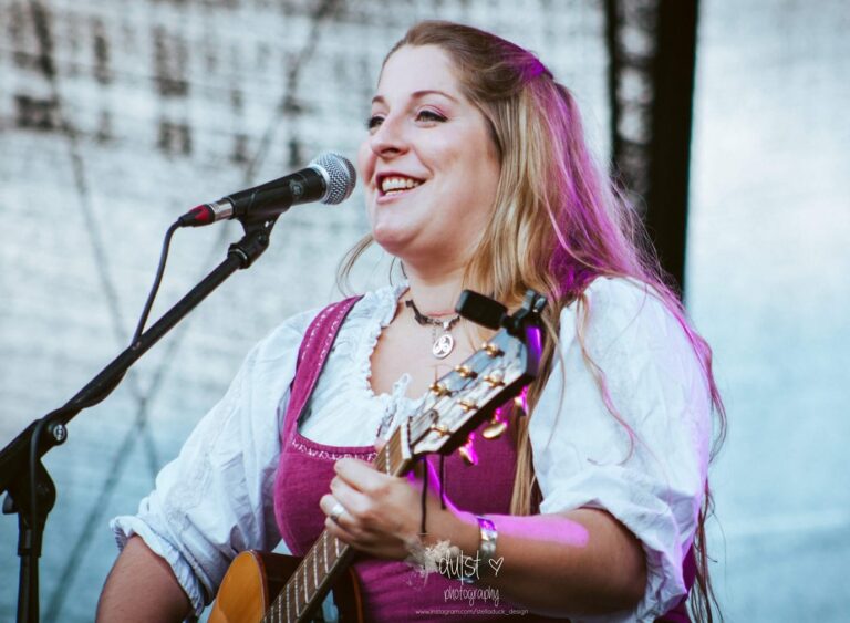 Frau mit langen blonden Haaren in traditioneller Trachtenbluse und pinkfarbener Weste, die lächelnd Gitarre spielt und vor einem Mikrofon auf einer Bühne steht.