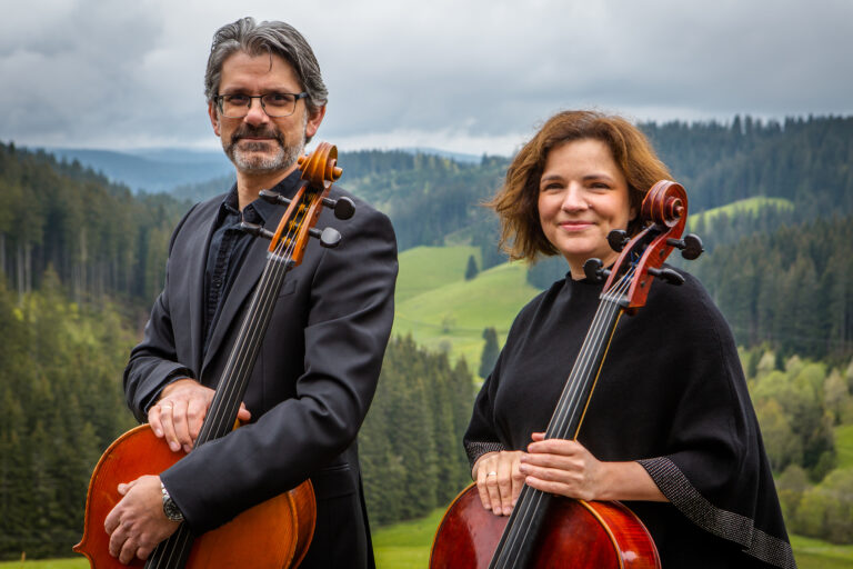 Zwei Cellisten, ein Mann mit Brille und grauem Haar und eine lächelnde Frau mit kurzen braunen Haaren, stehen in schwarzer Kleidung vor einer grünen, bewaldeten Berglandschaft mit bewölktem Himmel und halten ihre Celli.