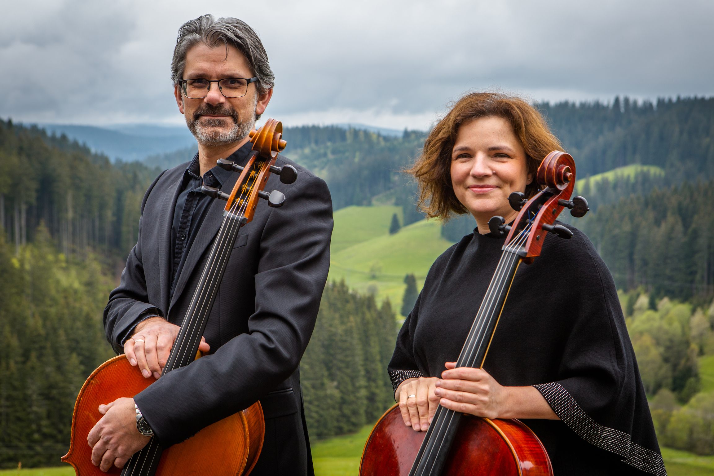 Zwei Cellisten, ein Mann mit Brille und grauem Haar und eine lächelnde Frau mit kurzen braunen Haaren, stehen in schwarzer Kleidung vor einer grünen, bewaldeten Berglandschaft mit bewölktem Himmel und halten ihre Celli.