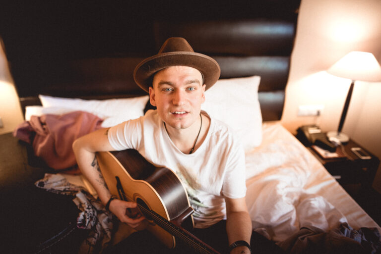 Junger Mann mit Hut, der auf einem Bett sitzt und eine akustische Gitarre spielt, umgeben von Kissen und einer Lampe auf einem Nachttisch.