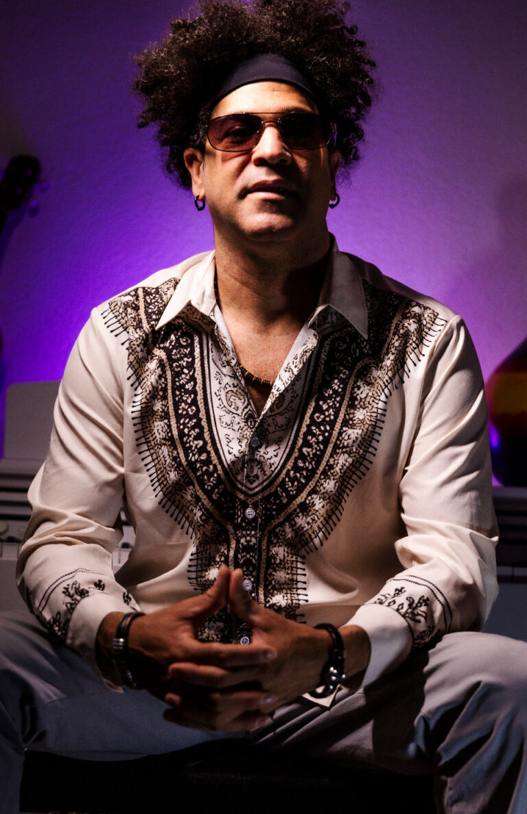 Porträt eines sitzenden Mannes mit lockigem Haar, Sonnenbrille und einem beigen Hemd mit braunem ethnischem Muster, vor einem violett beleuchteten Hintergrund mit einem Gitarrenhälfte sichtbar links