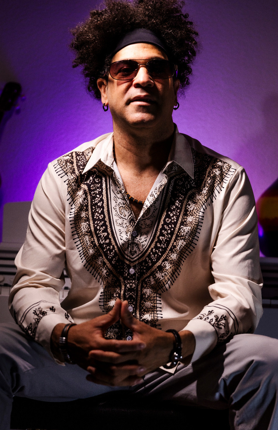 Porträt eines sitzenden Mannes mit lockigem Haar, Sonnenbrille und einem beigen Hemd mit braunem ethnischem Muster, vor einem violett beleuchteten Hintergrund mit einem Gitarrenhälfte sichtbar links