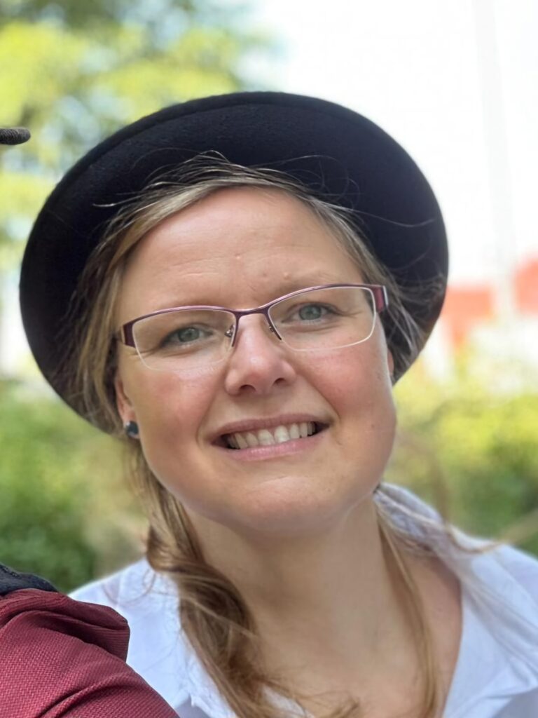 Porträt einer lächelnden Person mit Brille, schwarzen Hut und hellem Hemd vor unscharf grünem Hintergrund im Freien