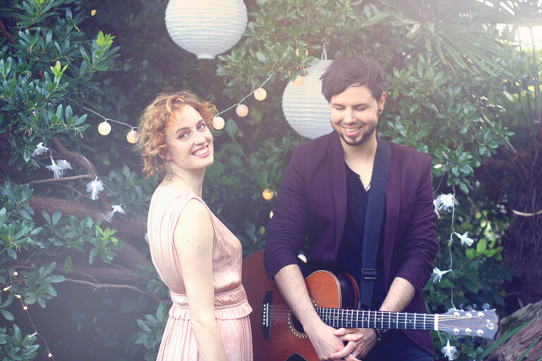 Frau in hellrosa Kleid und Mann mit dunkler Jacke, die Gitarre halten, vor beleuchteten Papierlaternen und grünem Laub, beide lächelnd in einer Gartenkulisse