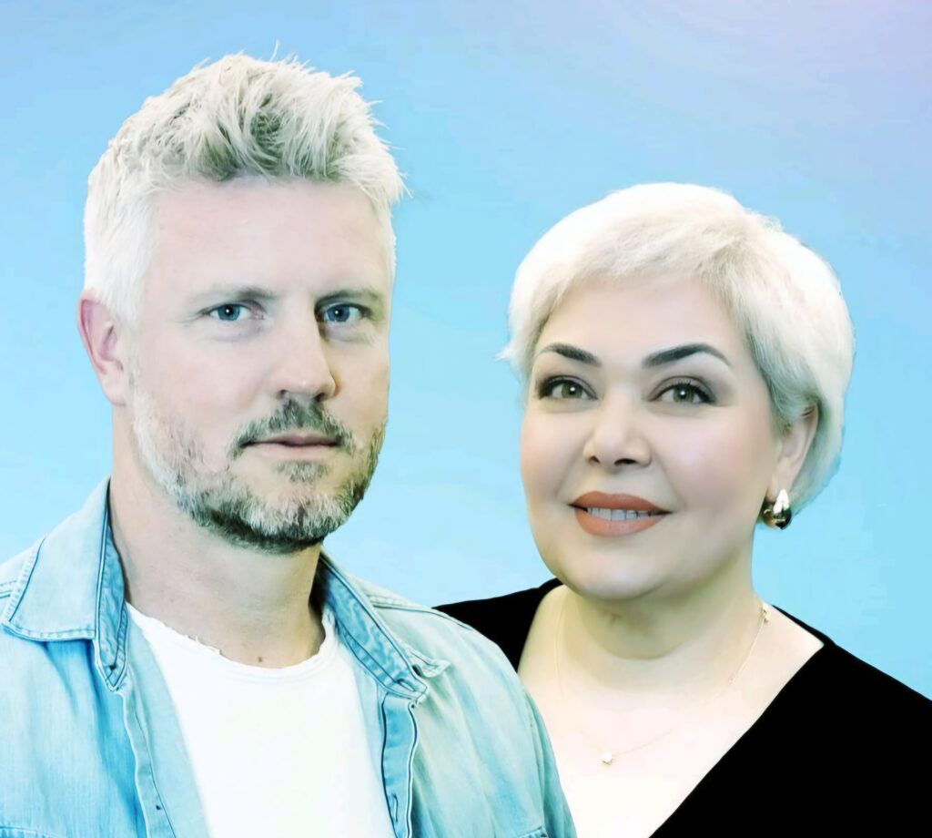 Porträt eines Mannes mit kurzen, hellblonden Haaren und Bart in Jeansjacke neben einer Frau mit kurzen weißen Haaren, roten Lippen und schwarzem Oberteil vor blauem Hintergrund