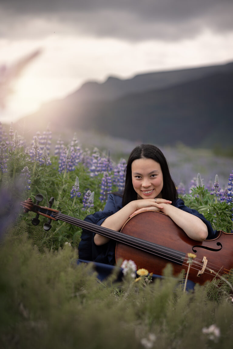Frau mit dunklen Haaren sitzt auf einer Wiese mit lila Blumen und umarmt ein Cello, während sie in die Kamera lächelt, im Hintergrund Berge und bewölkter Himmel mit Sonnenlicht.
