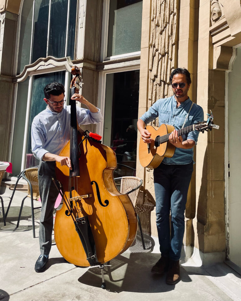 Zwei Männer spielen Musik auf einer sonnigen Terrasse vor einem Gebäude mit großen Fenstern, einer spielt Kontrabass und der andere Gitarre, beide tragen Brillen und lässige Kleidung.