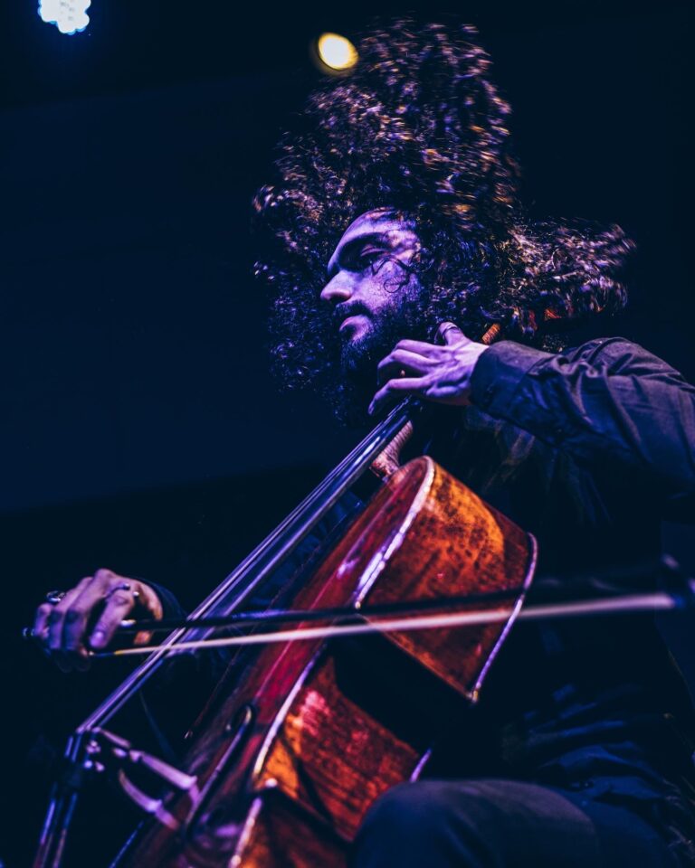 Ein Cellist mit langen, lockigen Haaren spielt leidenschaftlich Cello auf einer Bühne unter blauem Bühnenlicht.