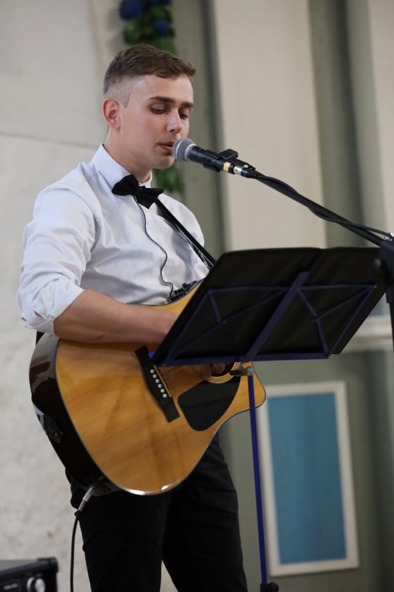 Junger Mann in weißem Hemd und schwarzer Fliege spielt akustische Gitarre und singt in ein Mikrofon vor einem Notenständer in Innenraum mit hellem Hintergrund.