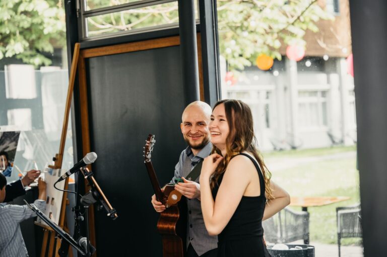 Ein Mann mit Glatze und Bart hält eine Akustikgitarre und lächelt, während eine Frau mit langen braunen Haaren und schwarzem Oberteil neben ihm lacht in einem hellen Raum mit Fenstern und Mikrofonständern im Vordergrund.