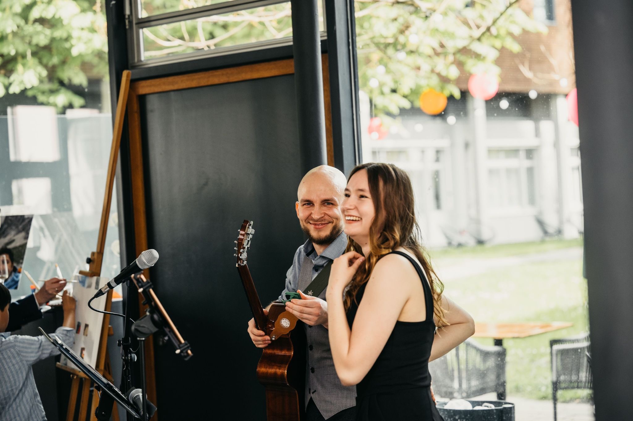 Ein Mann mit Glatze und Bart hält eine Akustikgitarre und lächelt, während eine Frau mit langen braunen Haaren und schwarzem Oberteil neben ihm lacht in einem hellen Raum mit Fenstern und Mikrofonständern im Vordergrund.