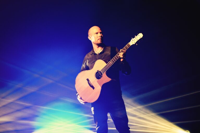 Mann mit Glatze und dunklem Hemd spielt Akustikgitarre auf Bühne mit blauem und gelbem Bühnenlicht im Hintergrund