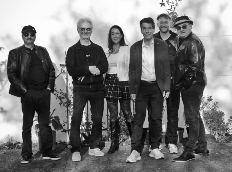 Schwarz-weiß-Foto von sechs Personen, fünf Männer und eine Frau, die vor einer hellen Wand stehen, zwei Männer tragen Lederjacken, die Frau trägt Rock, Stiefel und eine helle Jacke, die Gruppe steht im Freien auf einem Pflaster mit Pflanzen im Hintergrund.
