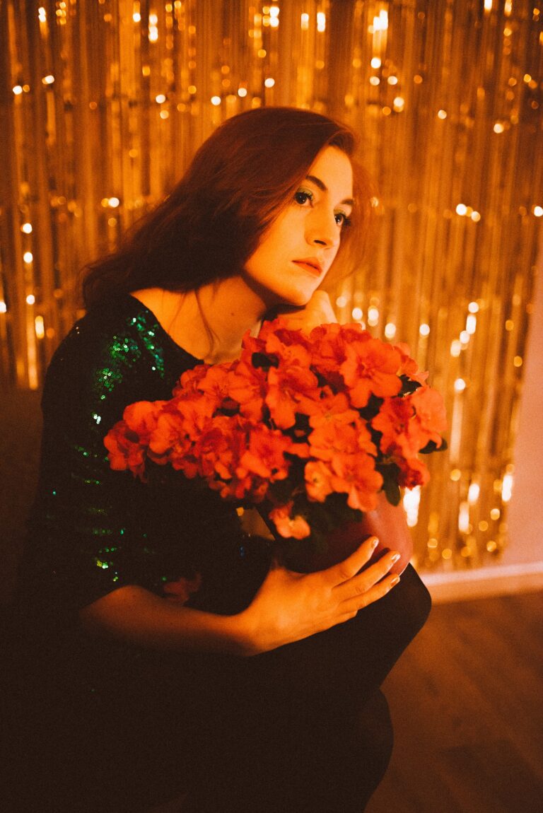 Frau mit langen braunen Haaren in grün glitzerndem Kleid, die nachdenklich einen großen Strauß roter Blumen hält, vor einem golden funkelnden Hintergrund sitzend