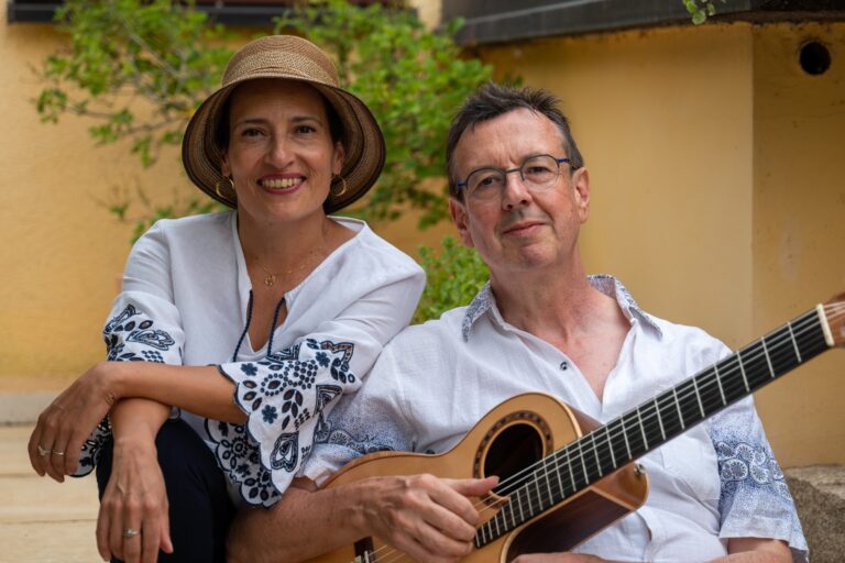 Zwei lächelnde Personen, eine Frau mit Strohhut und weißer Bluse mit blauen Mustern sitzt neben einem Mann mit Brille und weißem Hemd, der eine Gitarre hält, im Außenbereich vor gelber Wand und grünem Pflanzenhintergrund.