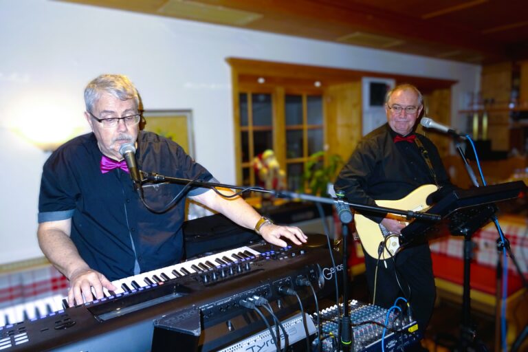Zwei ältere Männer musizieren in einem Raum, einer spielt Keyboard und singt in ein Mikrofon, der andere spielt E-Gitarre, beide tragen schwarze Hemden mit Fliegen, im Hintergrund Holzfenster und eine gemütliche Einrichtung.