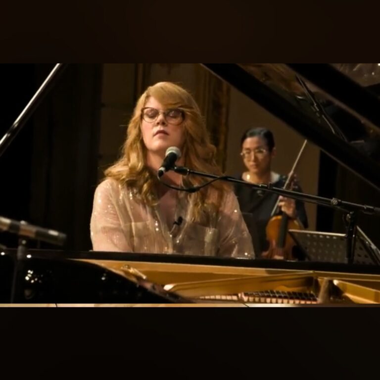 Frau mit blonden Haaren und Brille spielt Klavier und singt in ein Mikrofon, im Hintergrund hält eine Frau mit dunklen Haaren und Brille eine Violine, beide in einem Raum mit gedämpfter Beleuchtung.