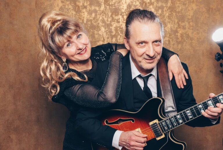 Ein Mann im schwarzen Anzug spielt eine E-Gitarre, während eine Frau mit langen blonden Haaren und einem schwarzen funkelnden Kleid ihn umarmt und beide lächeln vor einem goldbraunen Hintergrund.