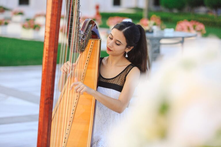 Junge Frau mit Pferdeschwanz und weißem Spitzenkleid spielt mit konzentriertem Blick auf einer Holzh arfe in einem Garten mit grüner Wiese und Blumen im Hintergrund