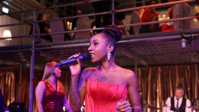 Frau mit elegantem roten Kleid und hochgestecktem Haar, die in einem Club oder einer Bar in ein Mikrofon singt, mit einer Band und einem Publikum im Hintergrund.