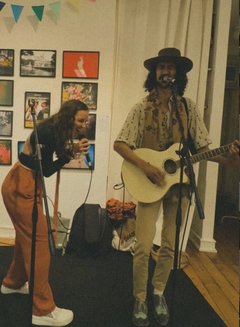 Eine Frau singt in ein Mikrofon, während ein Mann mit Hut und Schal neben ihr Gitarre spielt und singt, im Hintergrund bunte Bilder an einer Wand und bunte Wimpelleiste.
