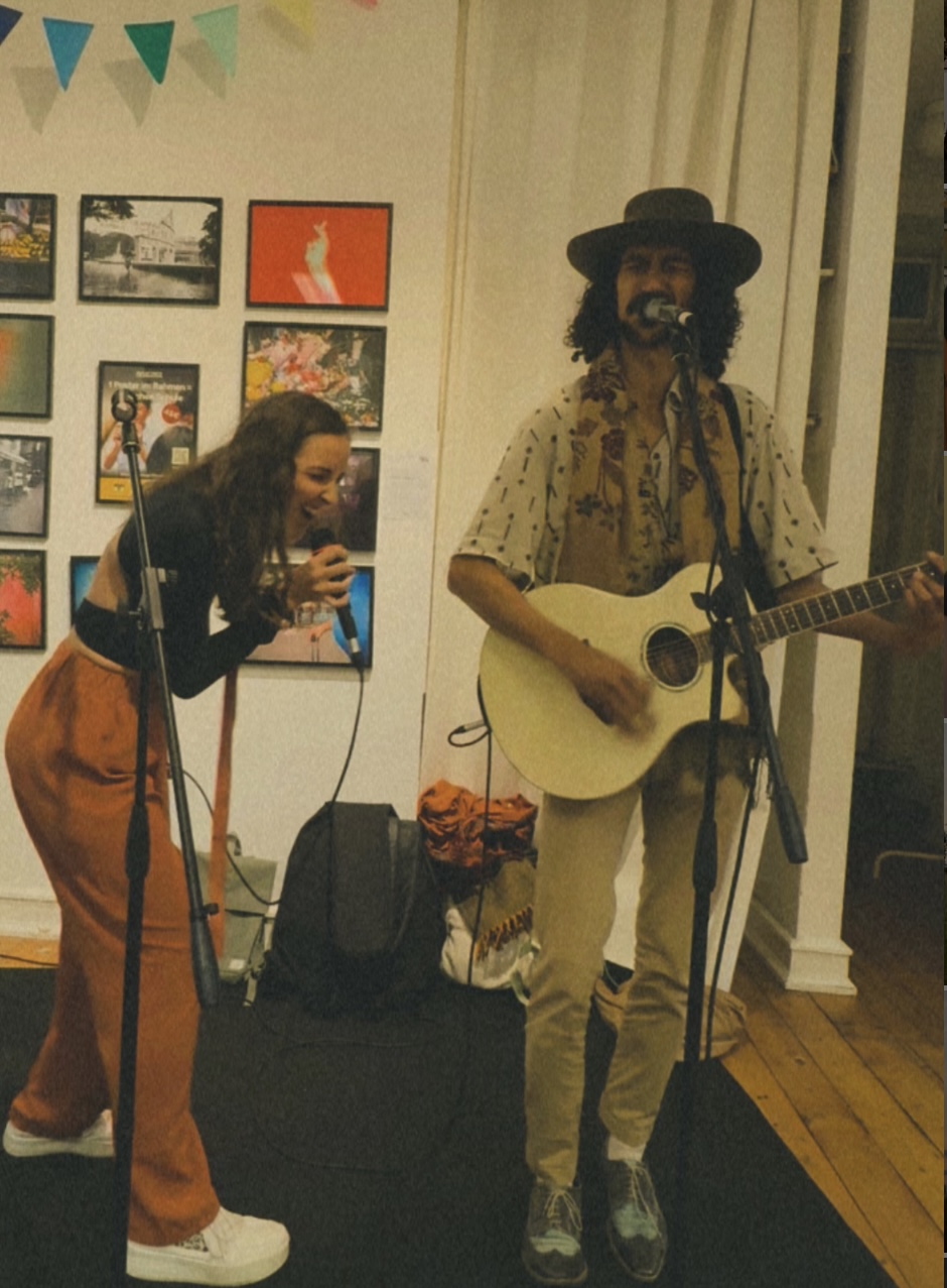 Eine Frau singt in ein Mikrofon, während ein Mann mit Hut und Schal neben ihr Gitarre spielt und singt, im Hintergrund bunte Bilder an einer Wand und bunte Wimpelleiste.