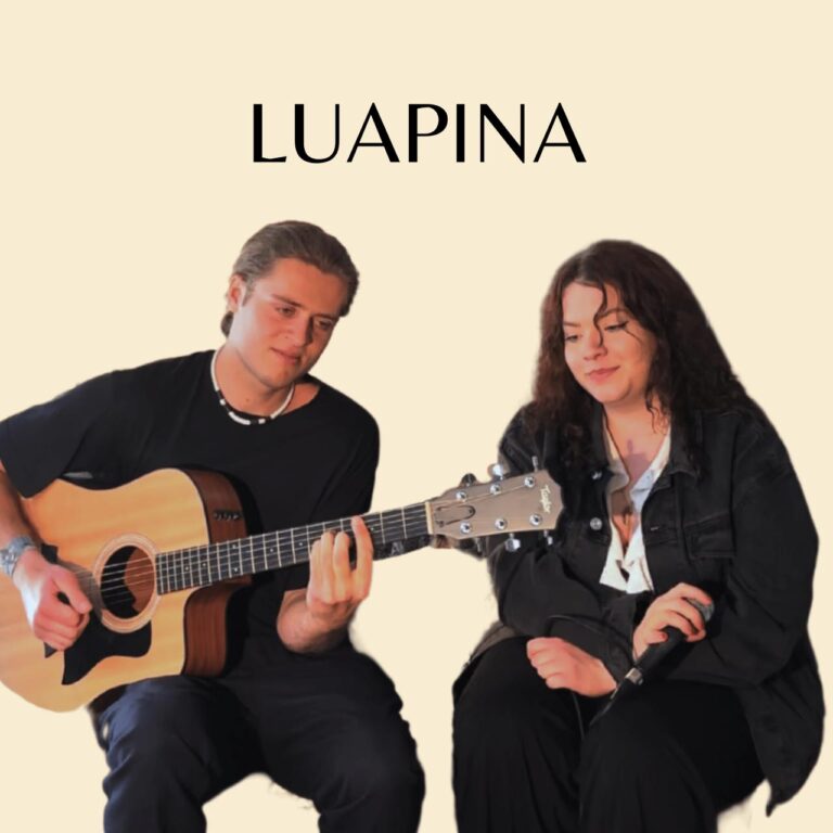 Zwei Personen sitzen vor hellem Hintergrund, links ein Mann mit akustischer Gitarre, rechts eine Frau, die ein Mikrofon hält, darüber Text mit dem Wort LUAPINA.