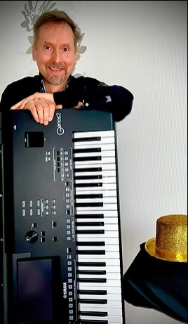 Mann mit Bart und kurzen Haaren lehnt lächelnd an einem Yamaha Keyboard, neben ihm steht ein goldglänzender Zylinderhut auf schwarzem Stoff.