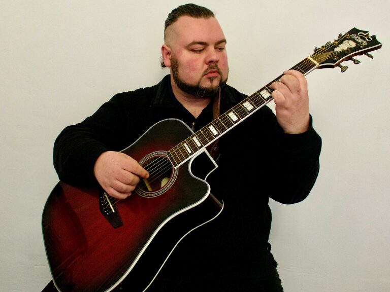 Mann mit schwarzen Haaren und Bart spielt Gitarre vor weißer Wand, trägt schwarzen Pullover und konzentriert sich auf das Gitarrenspiel.
